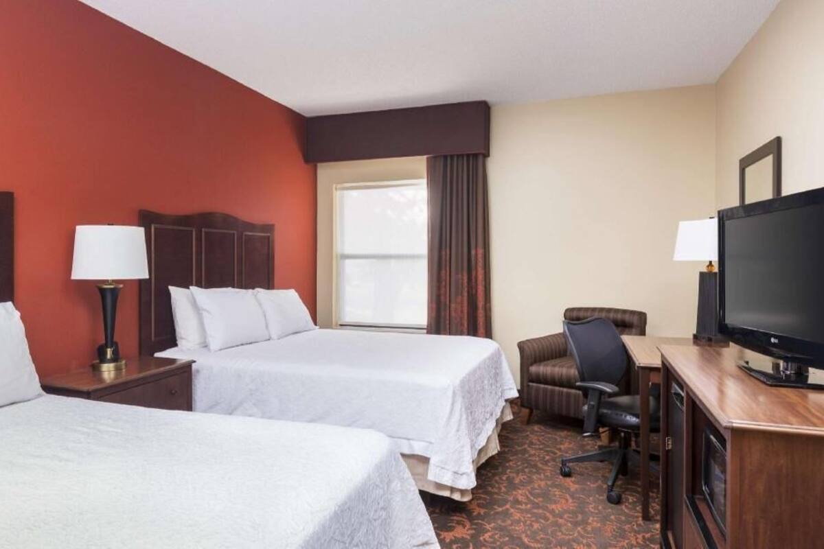 Habitación de hotel con dos camas, un escritorio con televisor, una silla y una ventana con cortinas.