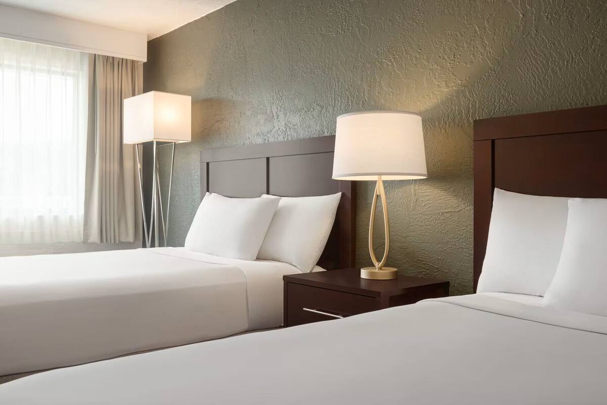 Habitación de hotel con dos camas, cabeceros, mesitas de noche, lámparas y un ventanal con cortinas.