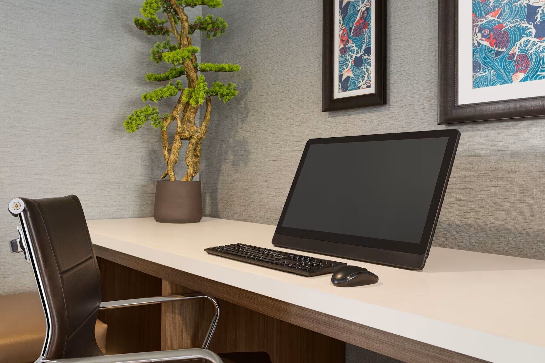 A modern office desk with a laptop, keyboard, and mouse, a potted bonsai tree, and framed artwork on the wall.
