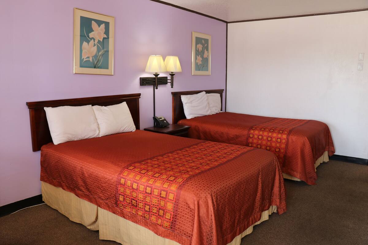 A hotel room with two beds, each with a red bedspread and white pillows. There are two wall-mounted lamps on a nightstand between the beds.