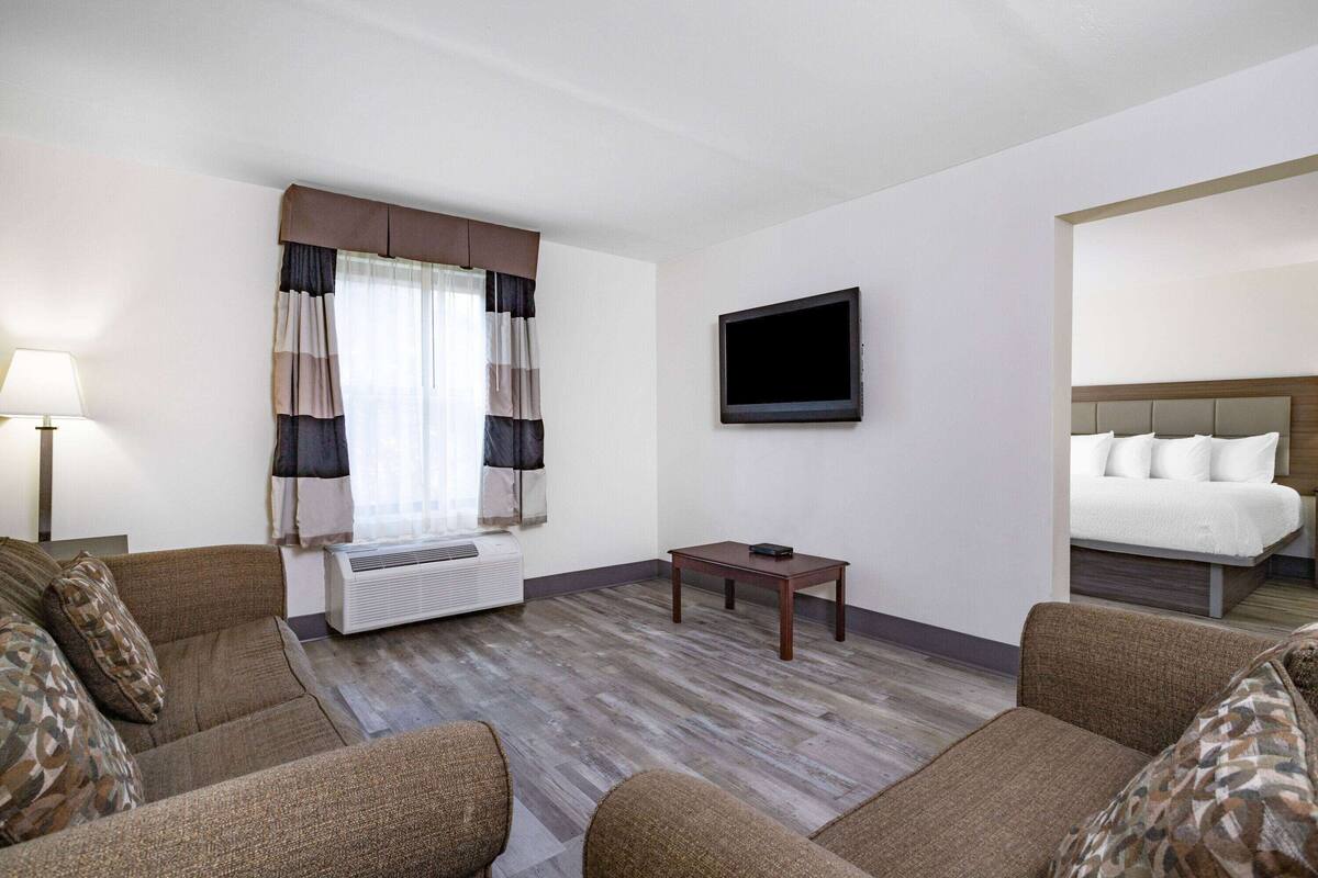 Habitación de hotel con sofá, una mesita, televisor de pantalla plana, cama con ropa blanca y una ventana con cortinas.