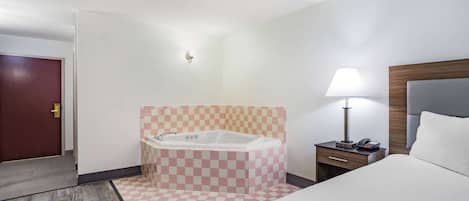 A hotel room with a checkered floor, a large bed, a nightstand with a lamp, and a bathroom with a checkered wall and a bathtub.