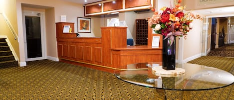 A reception area with a wooden reception desk, a glass table with a vase of flowers, patterned carpet, and a staircase.