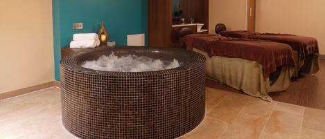 Un cuarto de spa con un jacuzzi, dos camillas de masaje y una zona de estar pequeña con un florero y toallas.