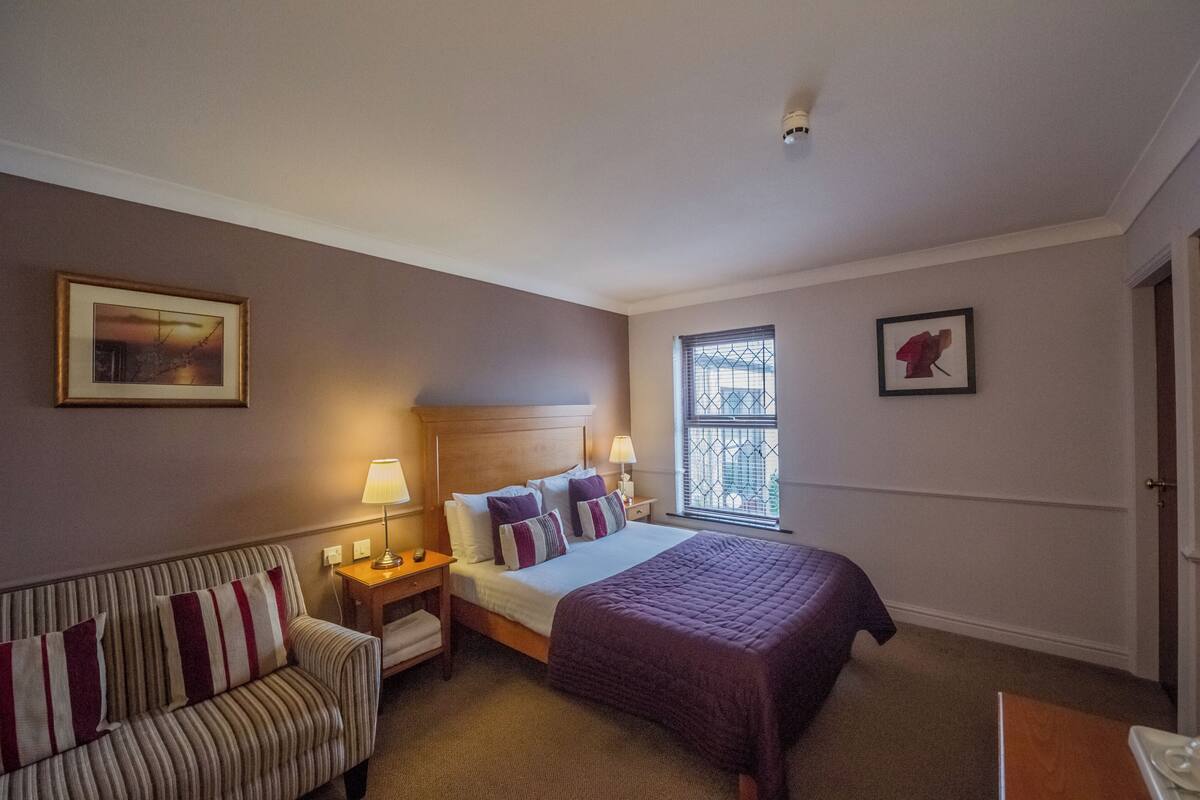 A hotel room with a bed, a sofa, a nightstand, two lamps, a window with a view, and two framed pictures on the wall.