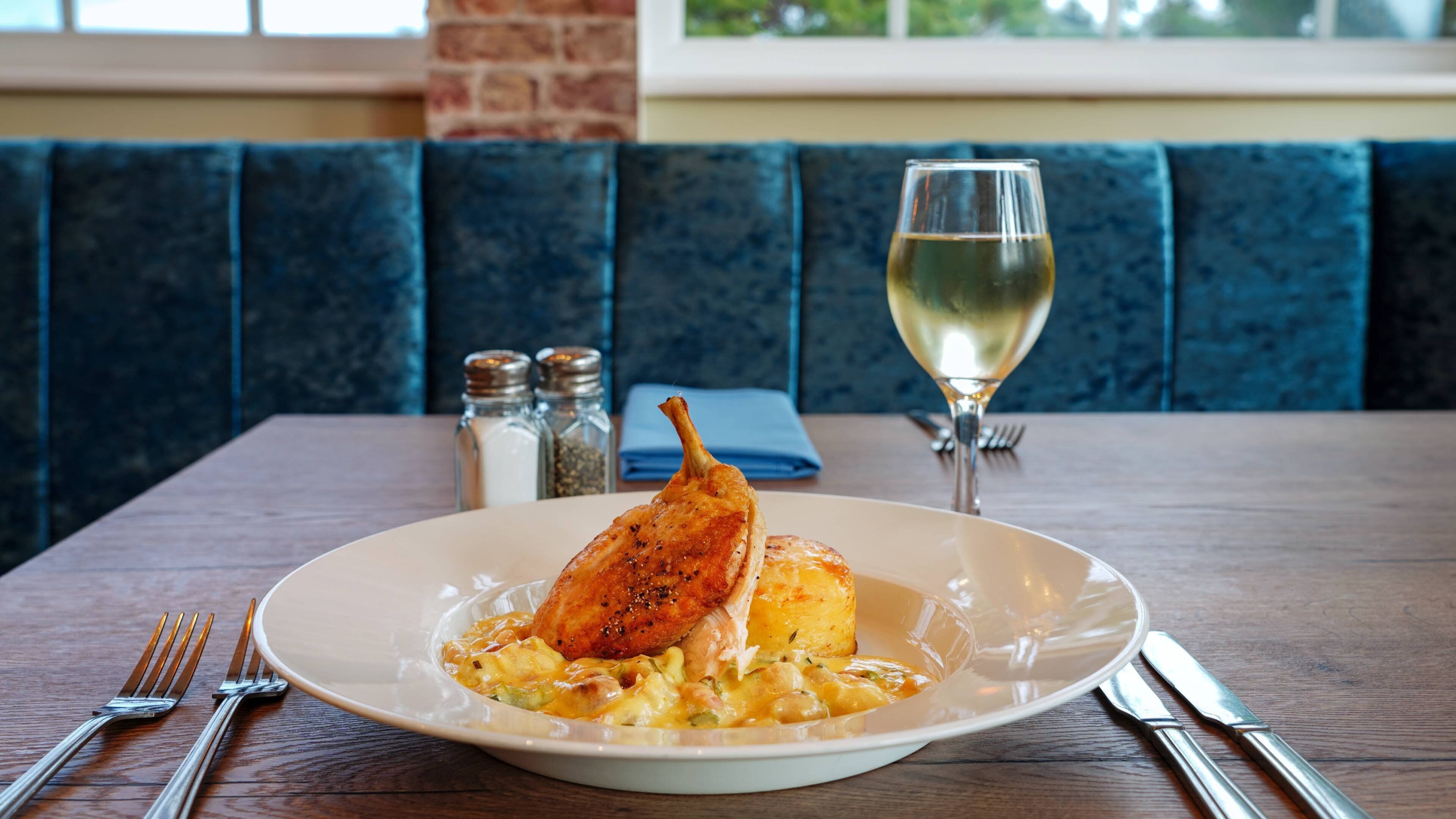 Une table à manger avec une assiette de nourriture, un verre de vin blanc et une banquette bleue rembourrée.