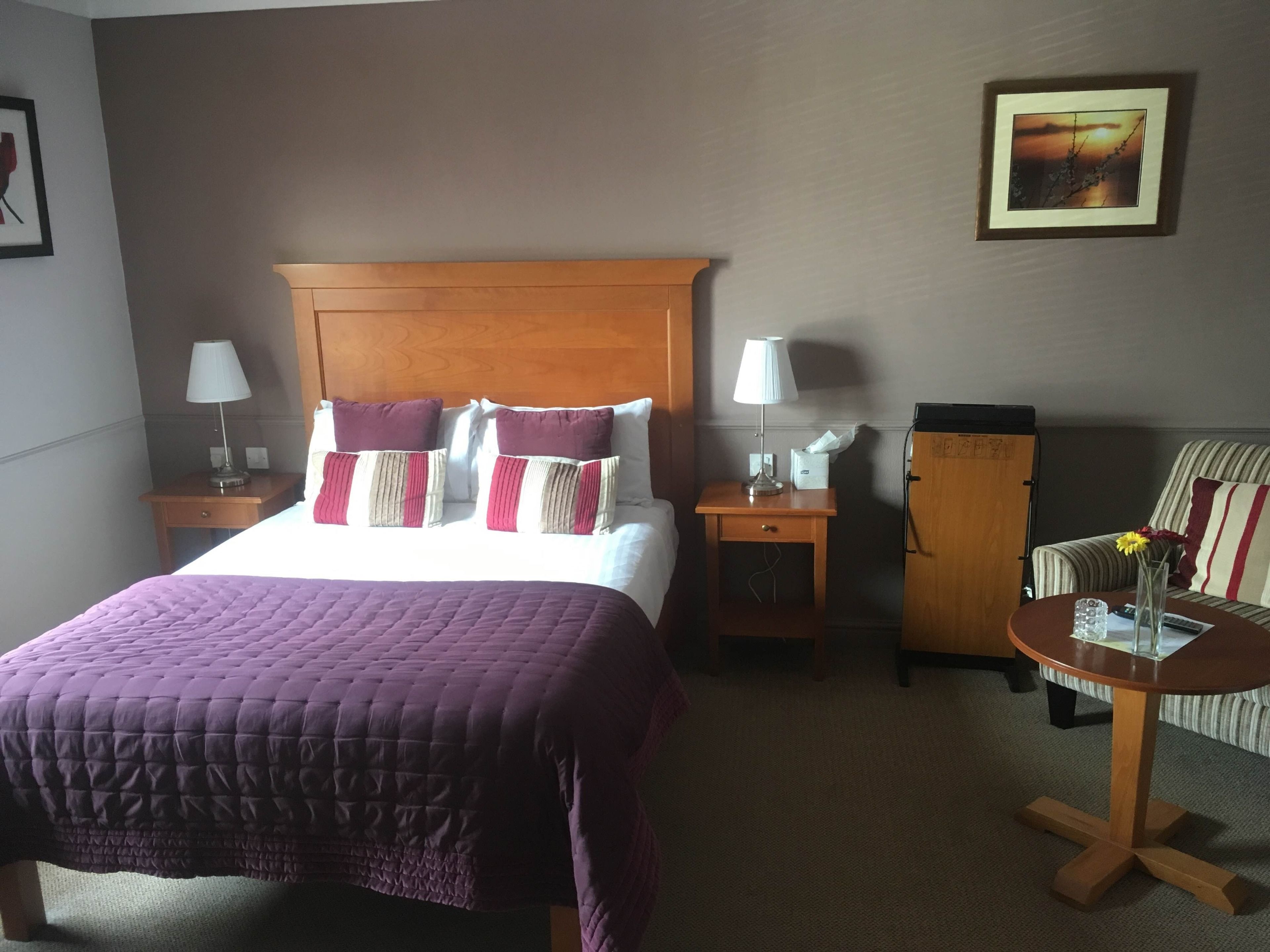 Habitación de hotel con una cama grande, dos mesitas de noche, una mesita auxiliar, una silla, una pequeña mesa con un florero y un cuadro en la pared.