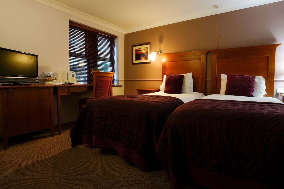 A hotel room with two beds, a desk with a television, and a window with blinds.