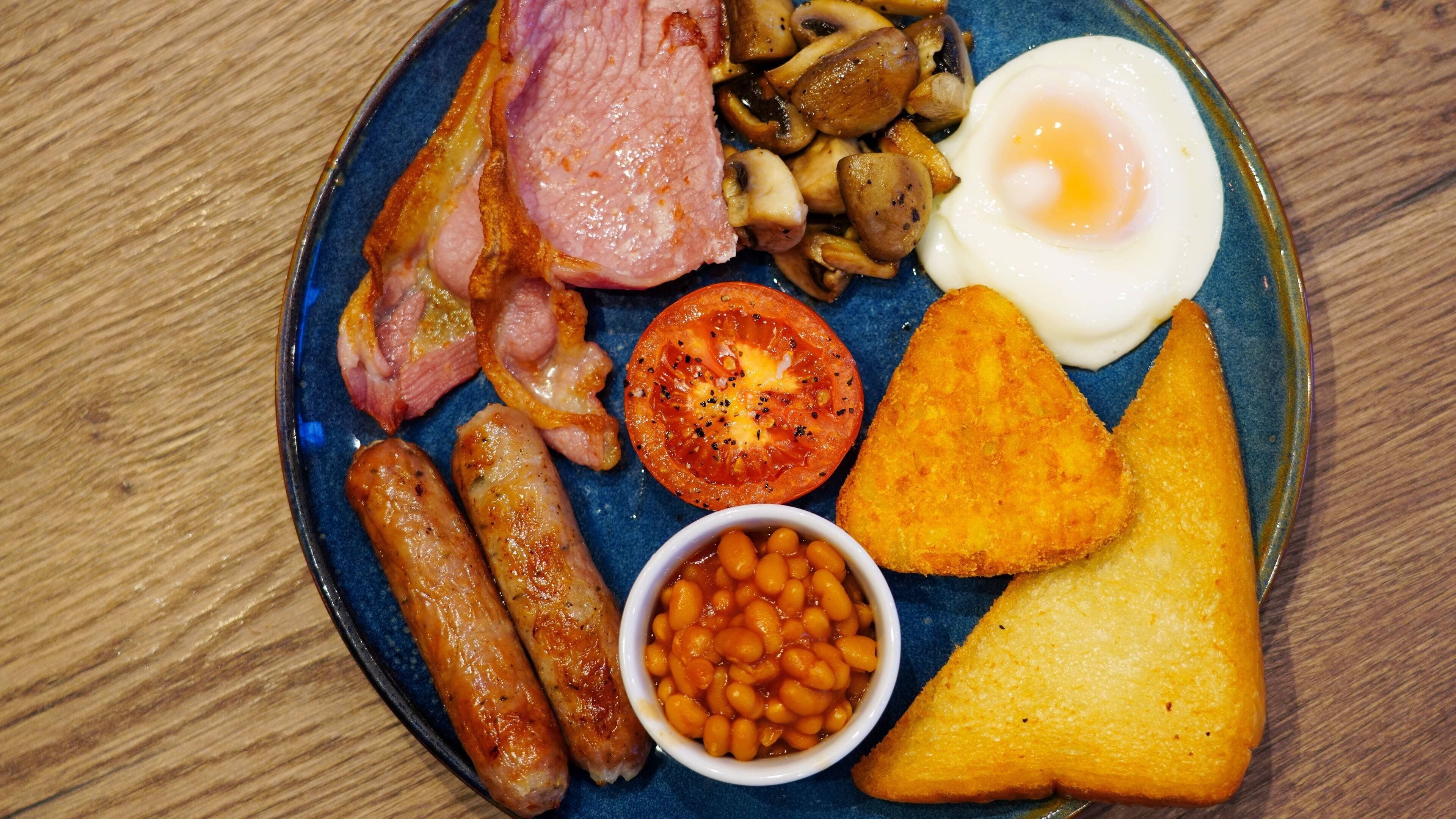 A plate of breakfast food including sausages, bacon, eggs, mushrooms, toast, and beans.