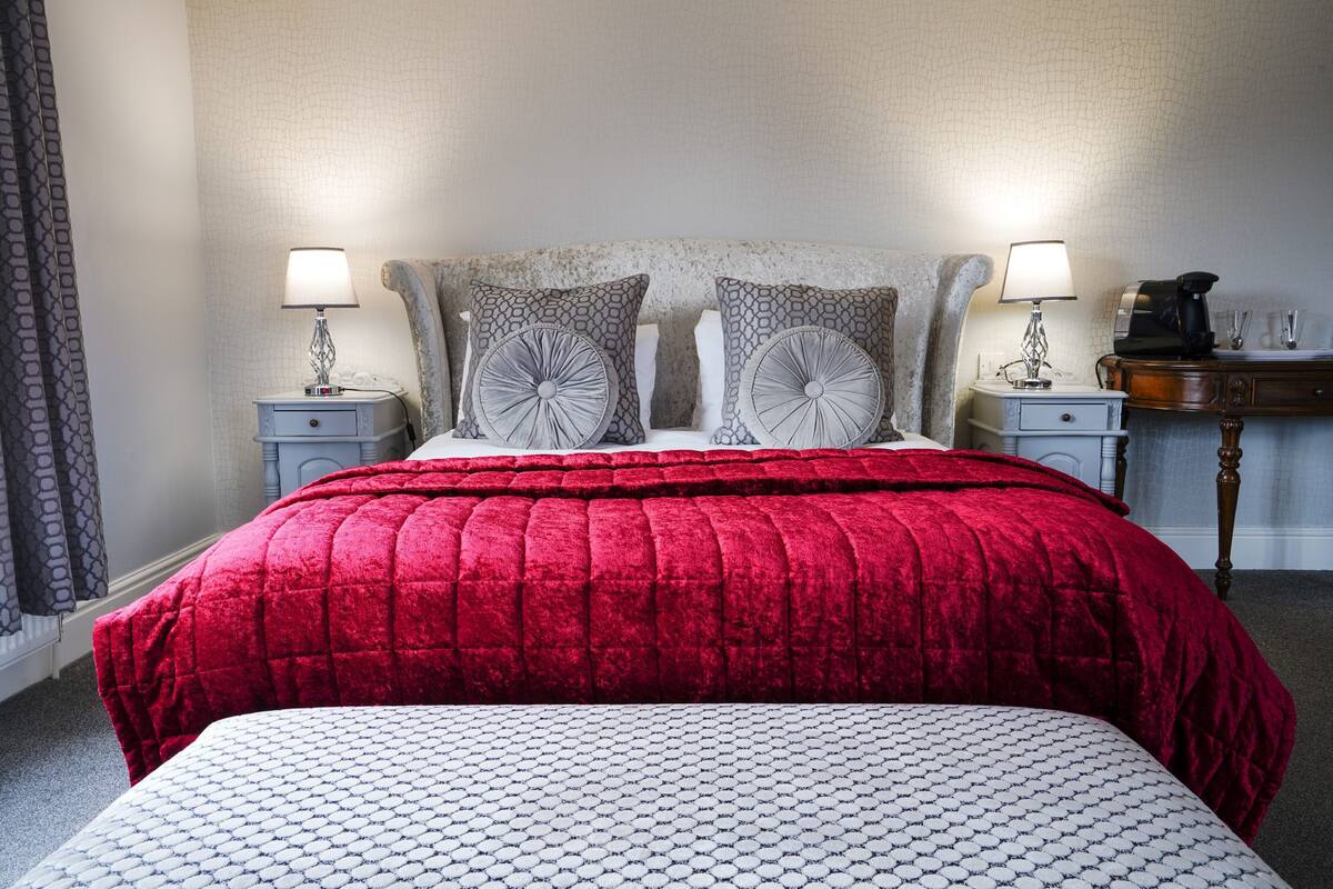 A hotel room with a large bed, two bedside tables, a nightstand, a coffee maker, and a desk with a lamp.