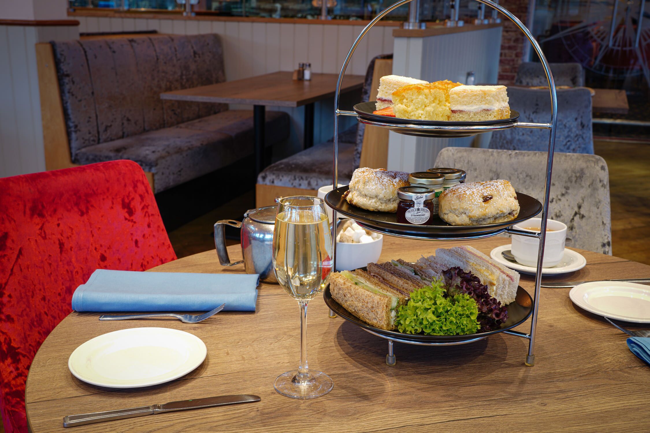 Un expositor de tres niveles con sándwiches y scones, una copa de champán y un mantelino azul sobre una mesa de madera en un restaurante.