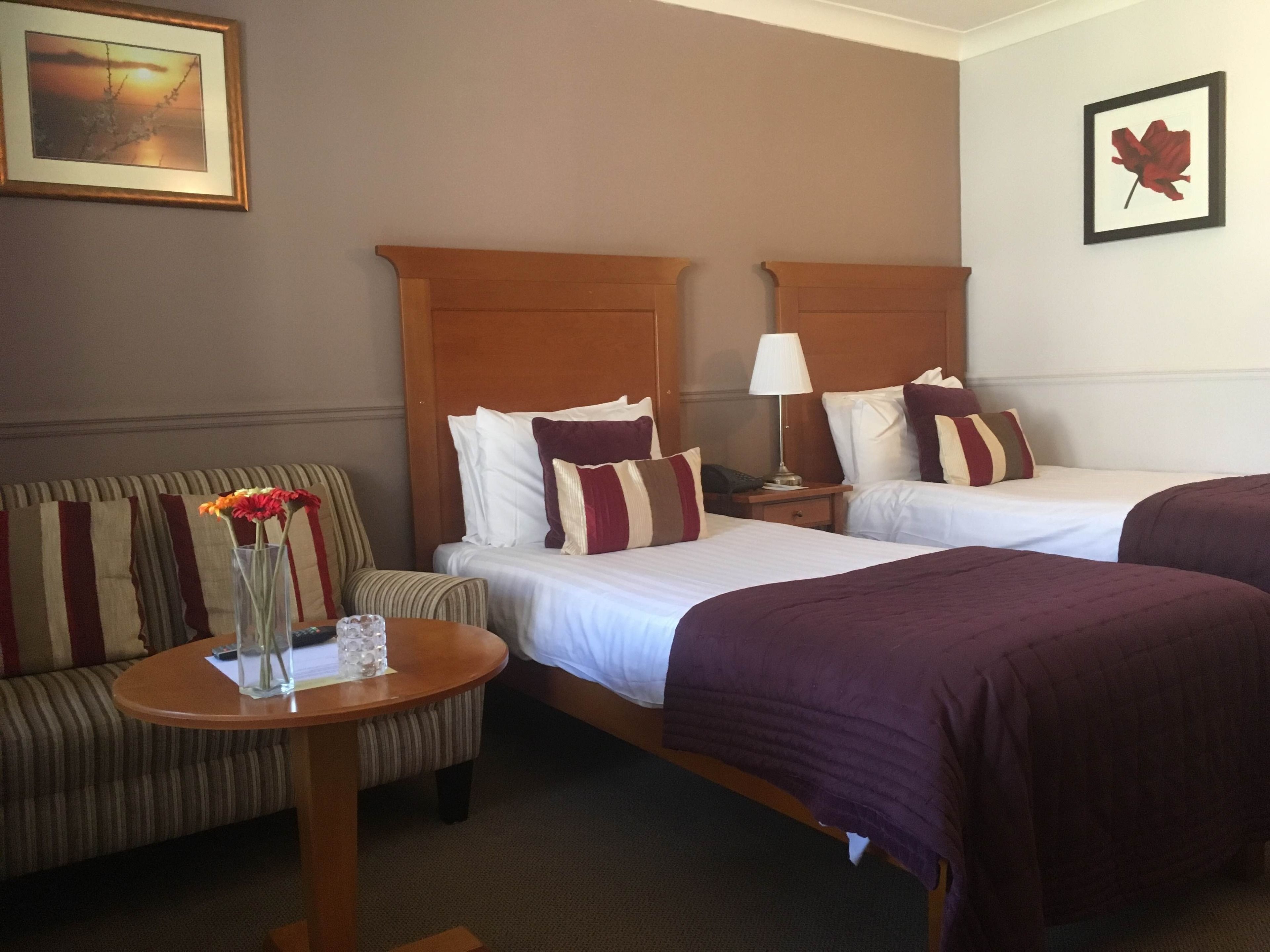 Habitación de hotel con dos camas, una mesita redonda pequeña con un florero, una silla y una mesita de noche con una lámpara.