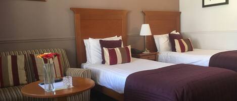 Habitación de hotel con dos camas, una mesita redonda pequeña con un florero, una silla y una mesita de noche con una lámpara.