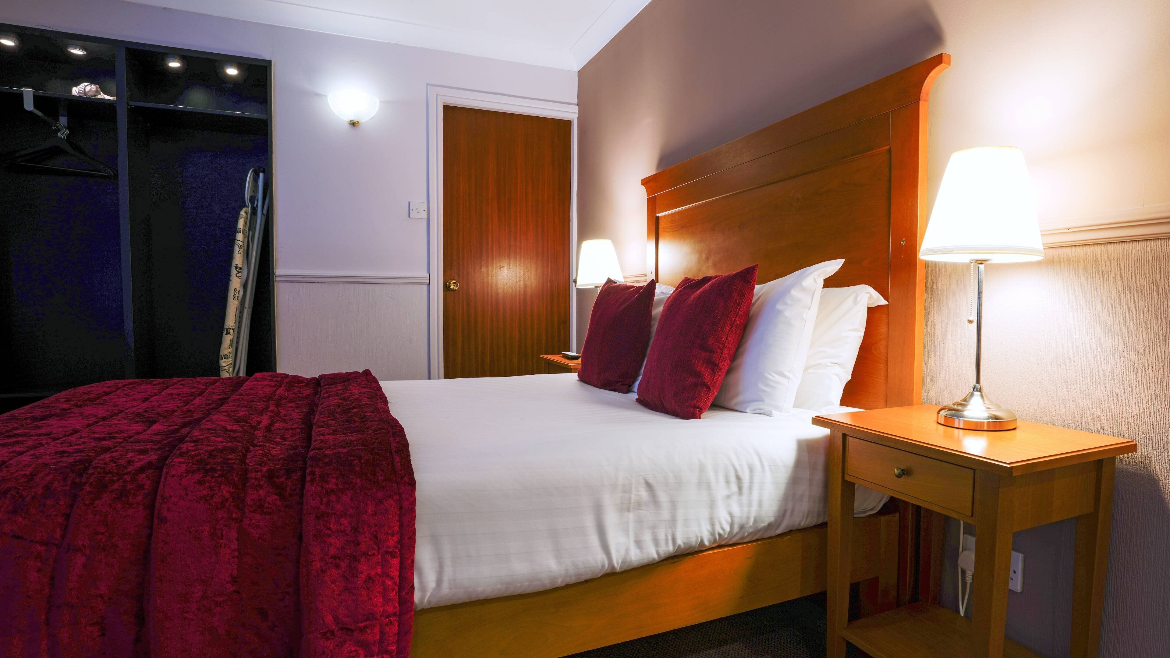 A hotel room with a bed, two bedside lamps, a wooden headboard, and a red throw blanket.