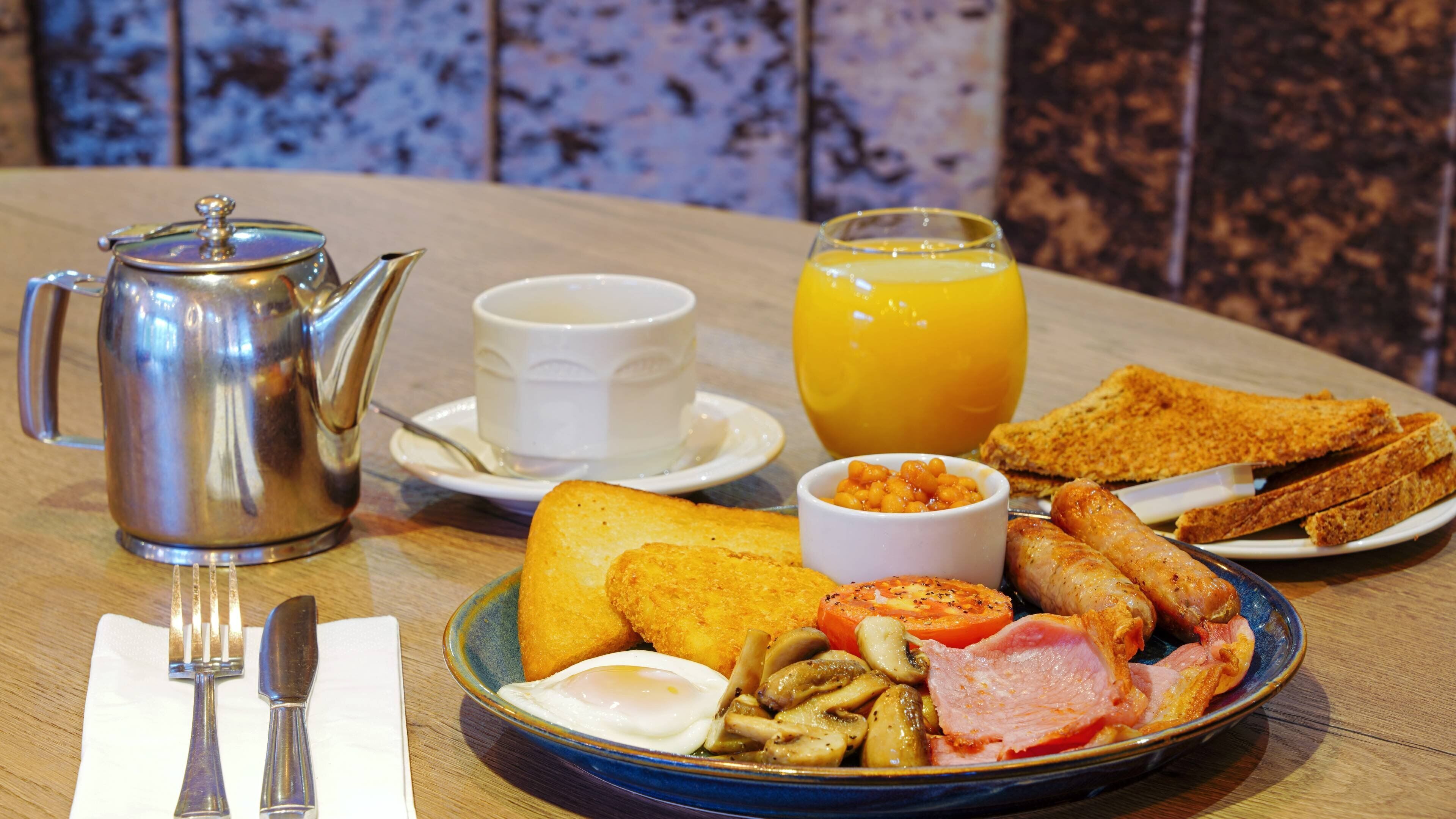 Ein Frühstückstisch mit Teekanne, Tasse, Orangensaft, Toast, Eiern, Pilzen und Schinken.