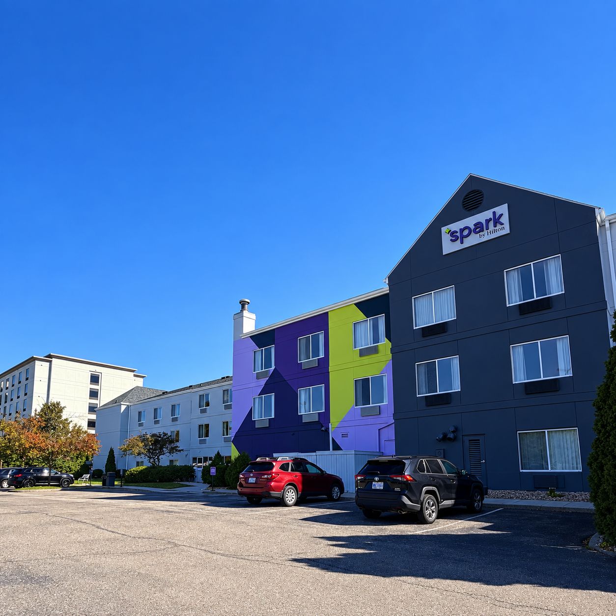 A multi-story hotel with a distinctive geometric facade, a parking lot with several cars, and a clear blue sky.