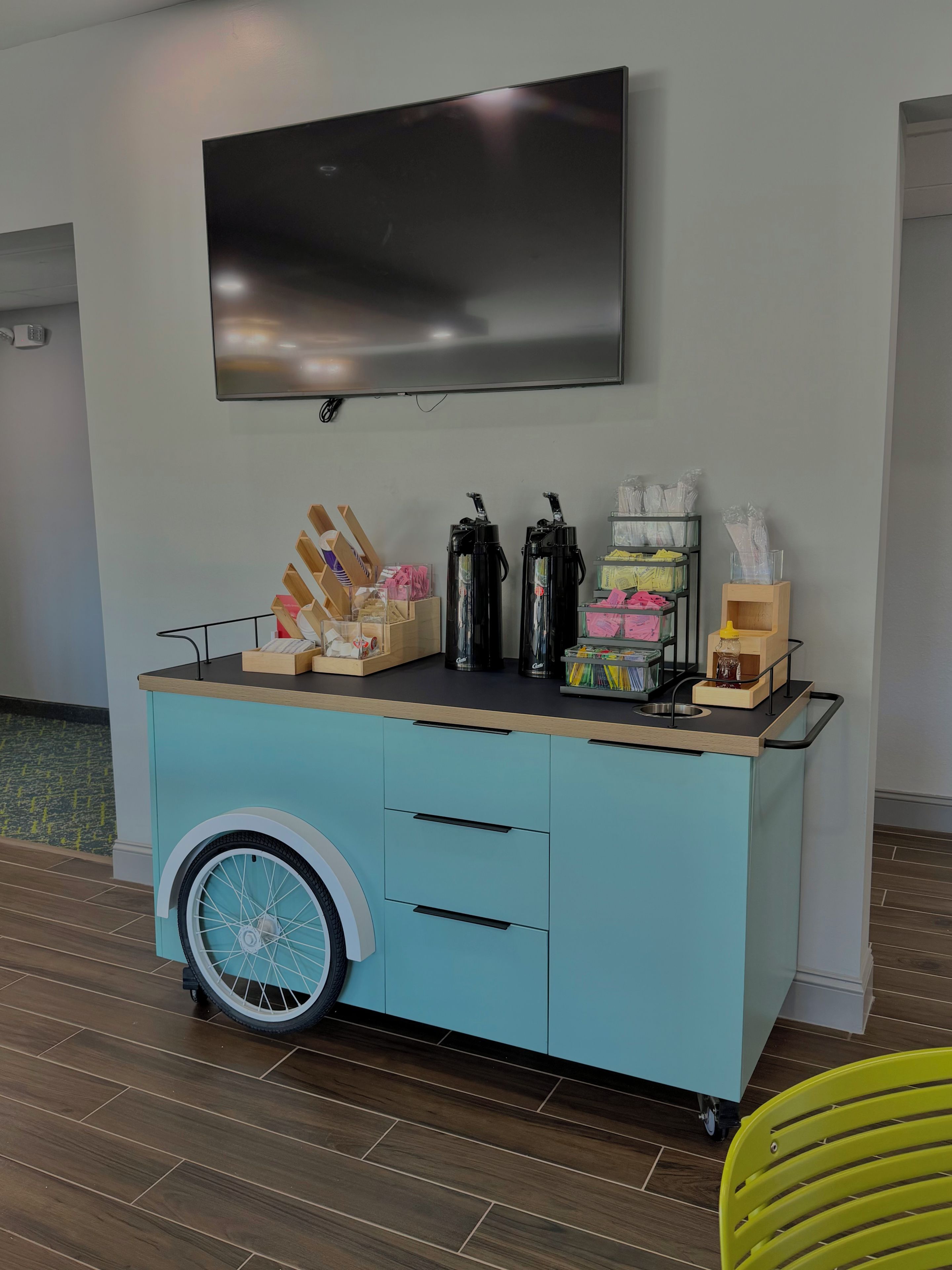 A teal-colored cart with a television mounted above it, a wheel, and various items on top including a coffee machine, a jar, and some boxes.