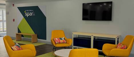 Un interior moderno con una zona de estar que incluye sillones mecedores amarillos y una mesa redonda, un televisor de pantalla plana en la pared, y una alfombra estampada color verde en el suelo.