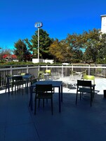 An outdoor seating area with tables and chairs, a white fence, and trees in the background.