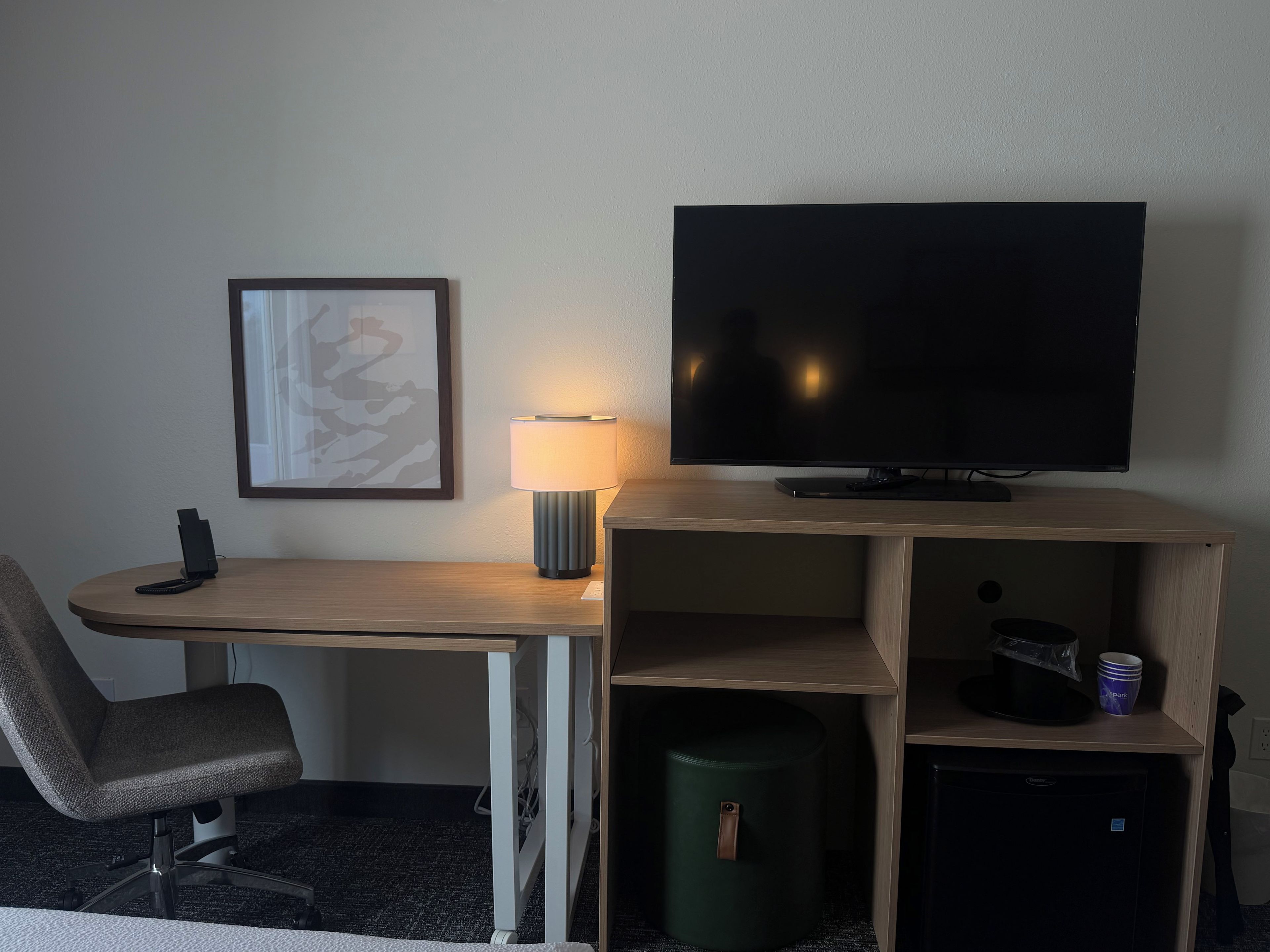 A hotel room with a desk, chair, TV, lamp, and a trash bin.