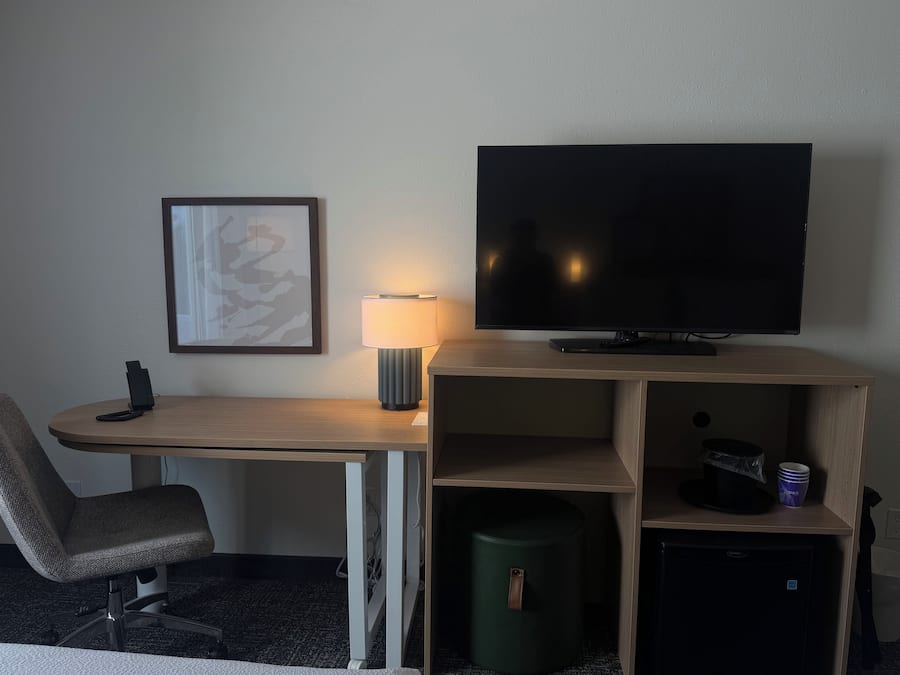 A hotel room with a desk, chair, TV, lamp, and a trash bin.