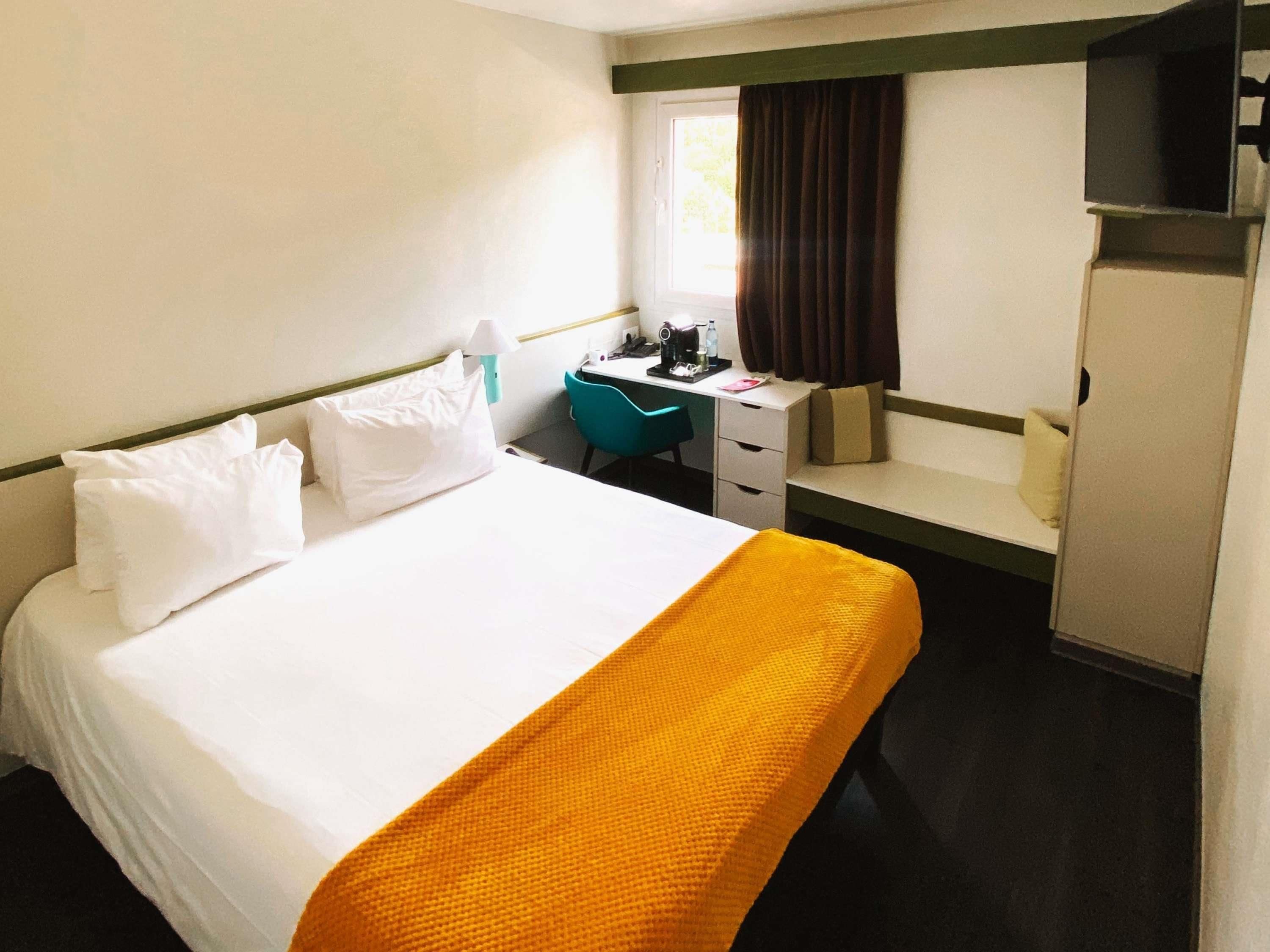 Habitación de hotel con cama, escritorio, silla, armario y ventana con cortinas.