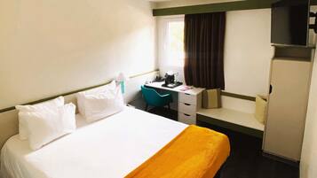 Habitación de hotel con cama, escritorio, silla, armario y ventana con cortinas.