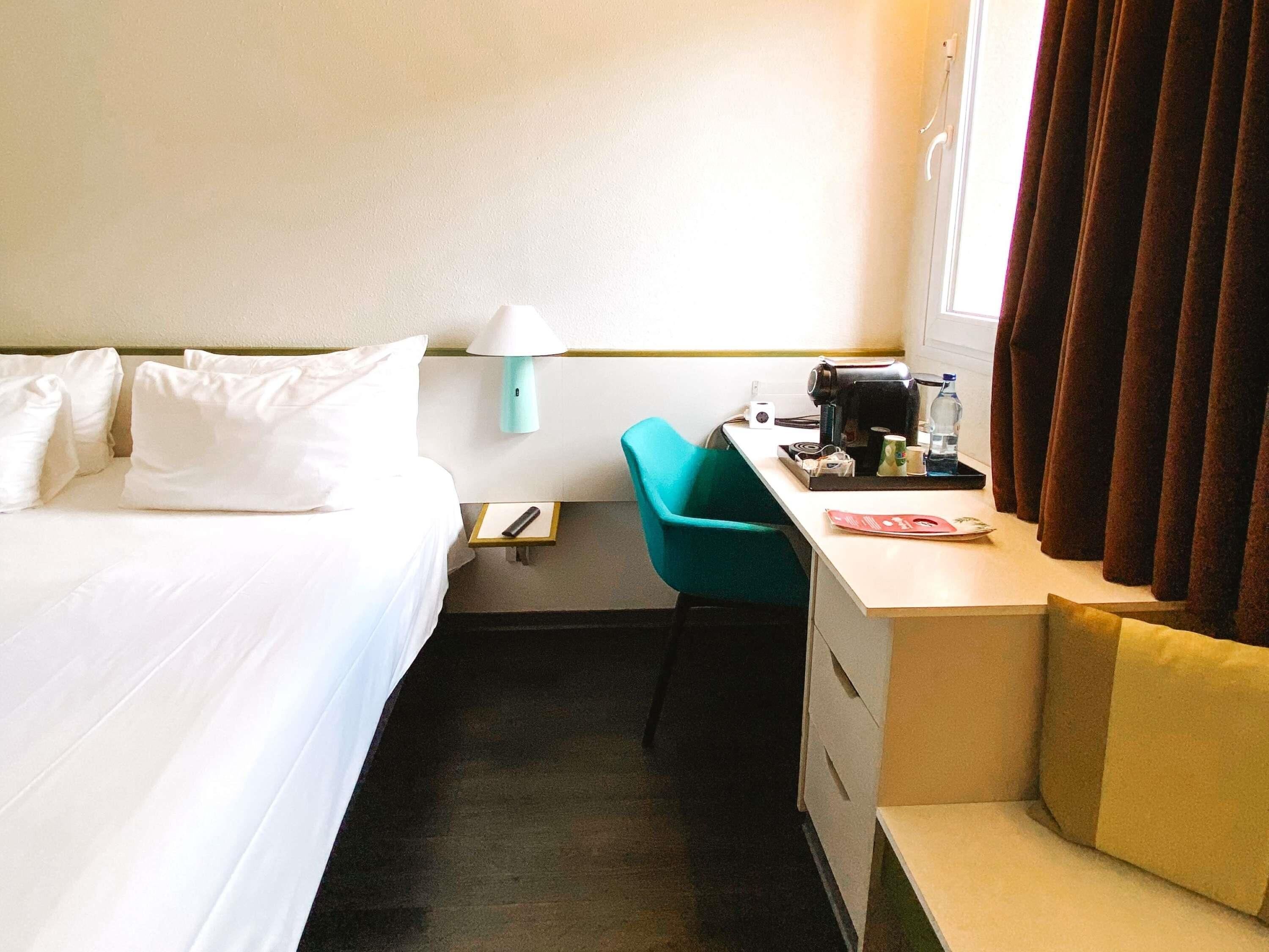 Una habitación de hotel con cama, escritorio, silla, lámpara y ventana con cortinas.