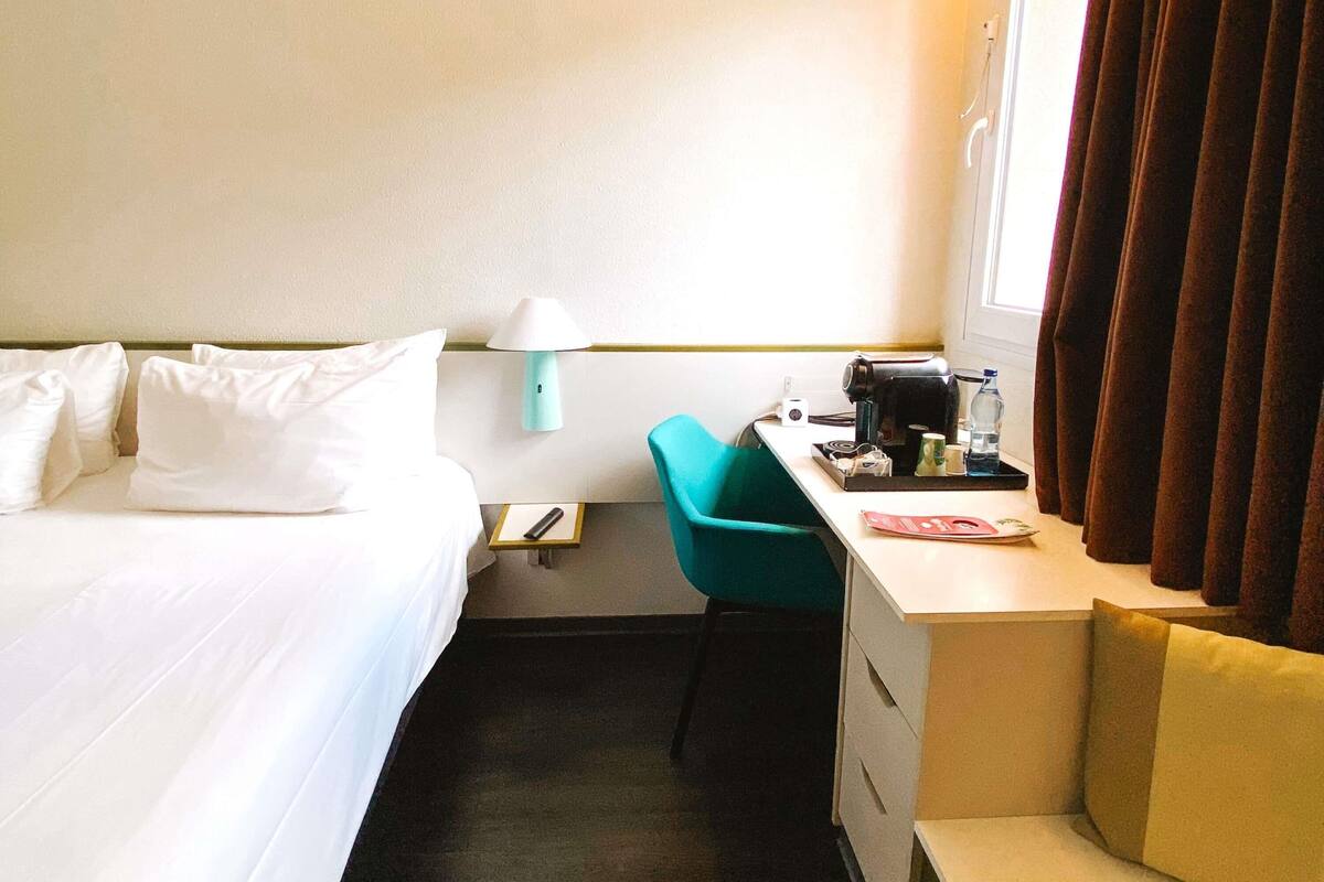 A hotel room with a bed, a desk with a chair, a lamp, a coffee maker, and a window with curtains.