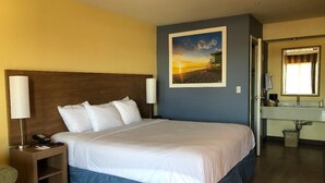 A hotel room with a large bed, a nightstand with a lamp, a bathroom with a sink and mirror, and a painting of a sunset on the wall.