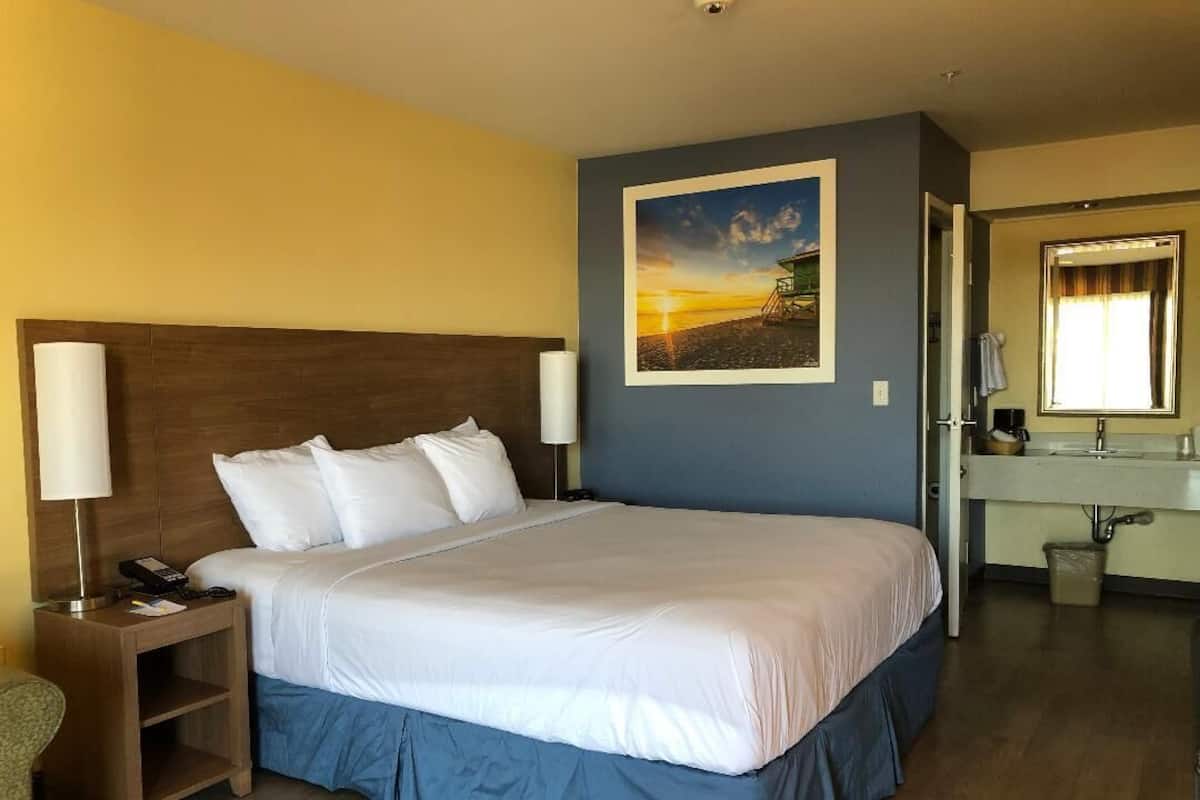 A hotel room with a large bed, a nightstand with a lamp, a bathroom with a sink and mirror, and a painting of a sunset on the wall.