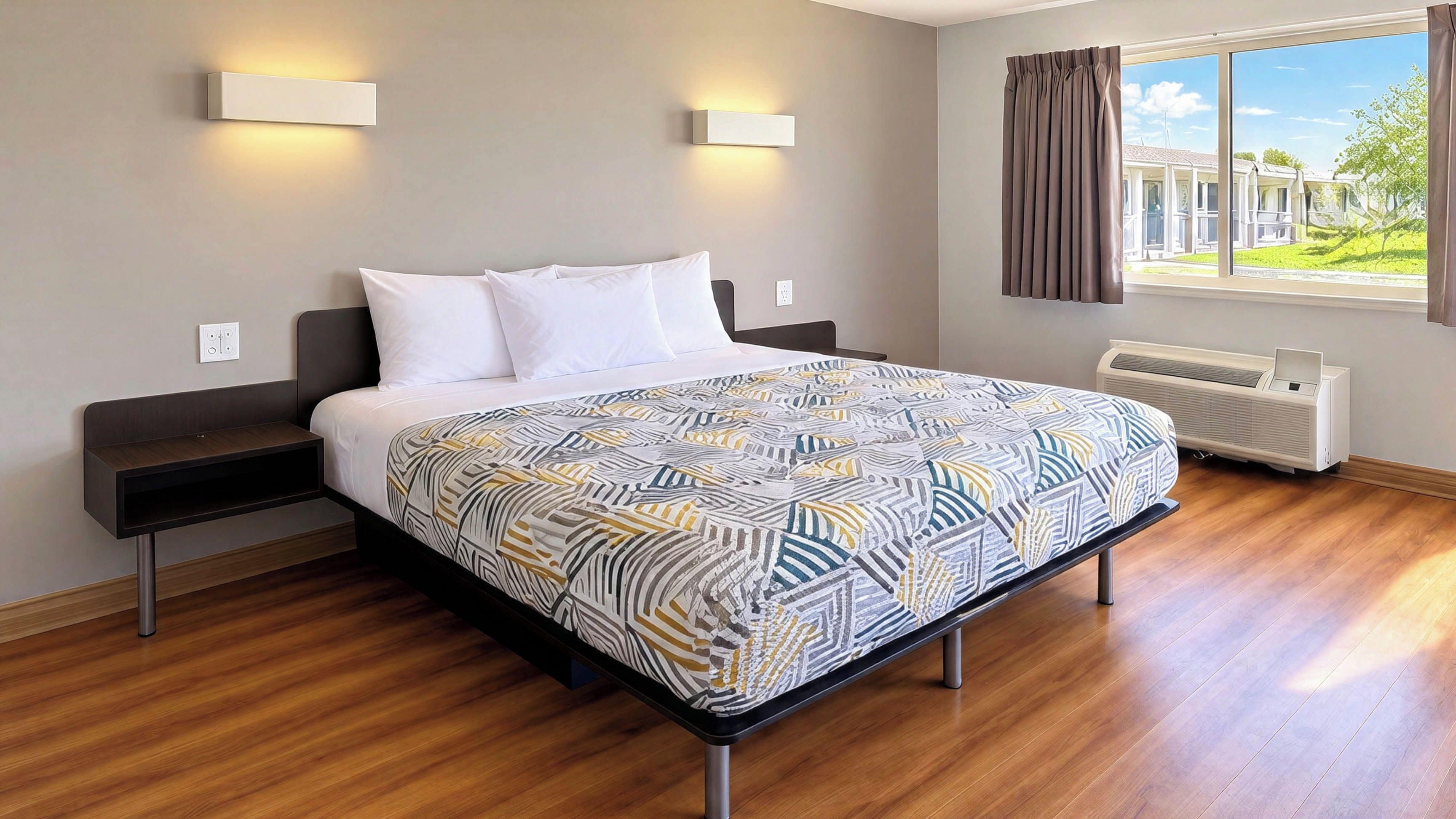 A hotel room with a bed, bedside tables, a window with curtains, and a wall-mounted air conditioner.