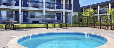 A swimming pool with a surrounding fence, a building with multiple balconies, and a small table with benches.