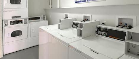 A laundry area with multiple washing machines and dryers, a control panel, and a wall-mounted information board.