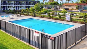 Una piscina rectangular con una cerca negra, rodeada de césped y un edificio de varios pisos al fondo.