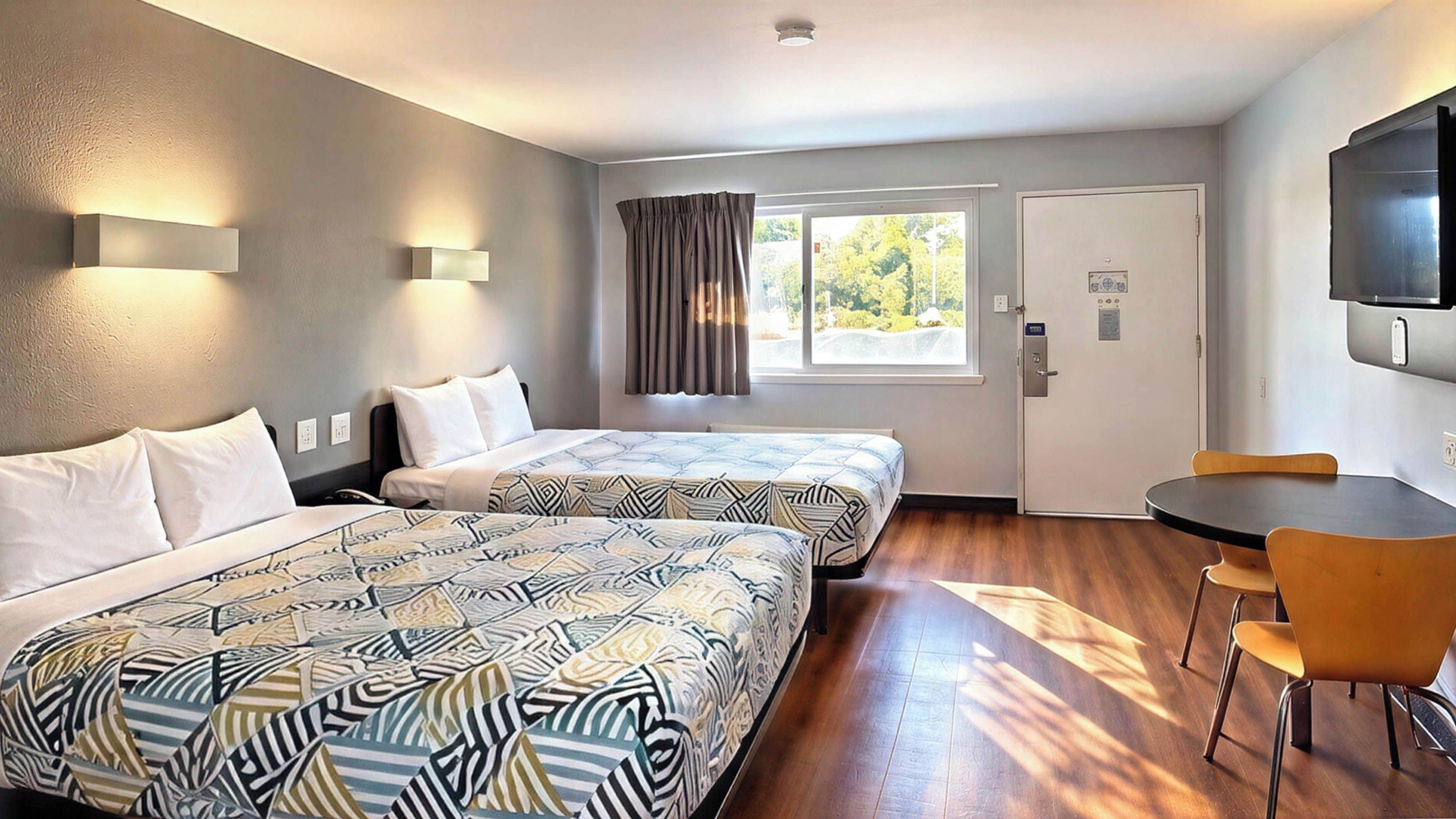 A hotel room with two beds, a dining table, chairs, a TV mounted on the wall, and a window with curtains.