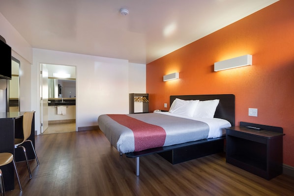 A modern hotel room with a bed, bedside table, and a small desk area. - Motel 6 Claremont, CA (Claremont)