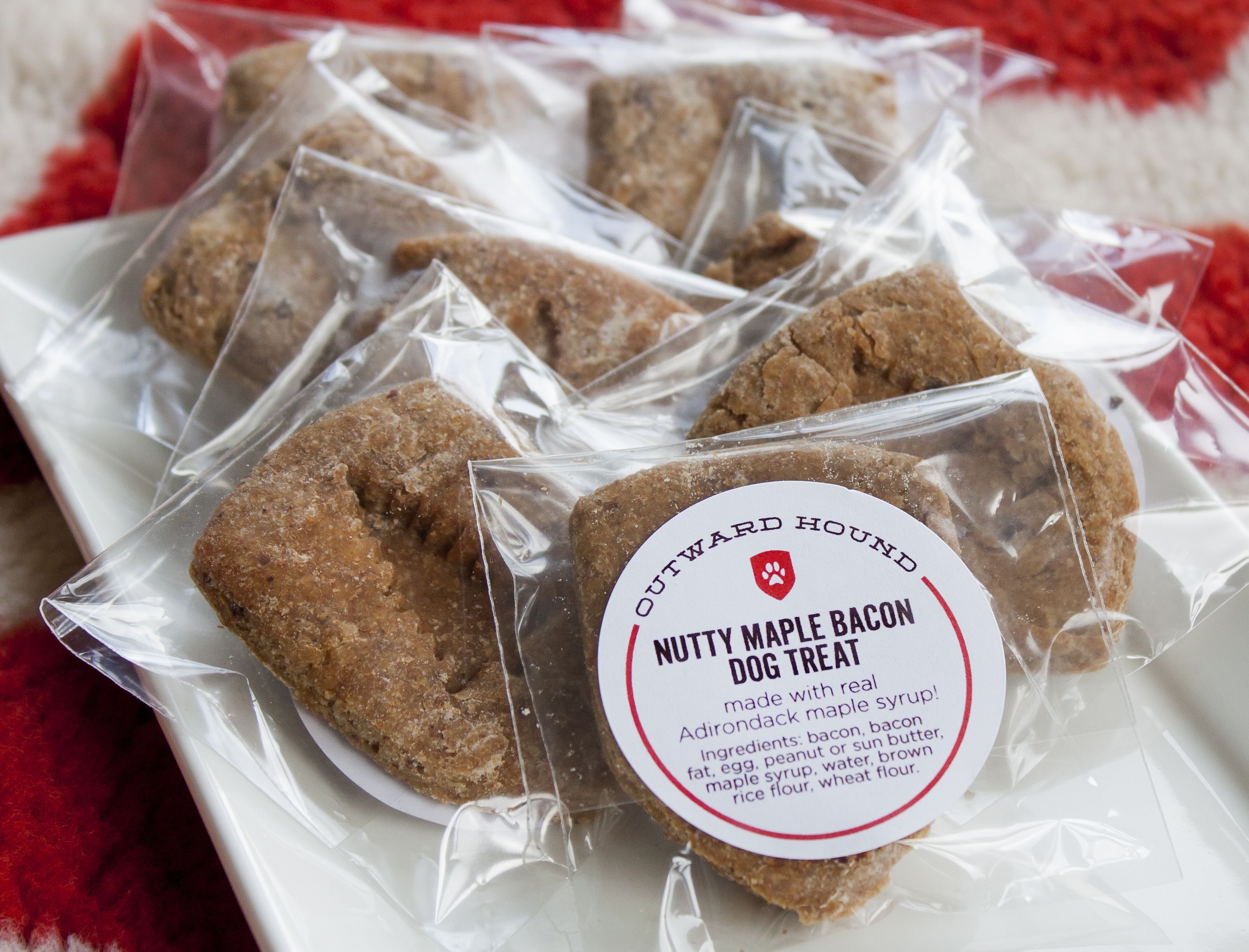 Ein Teller mit verpackten Hundeleckerlis, beschriftet mit 'Nutty Maple Bacon Dog Treat'. Die Zutatenliste enthält Speck, Ei, Erdnussbutter, Fett, Ei, Speck, Ahornsirup, Wasser, braunen Reismehl und Weizenmehl.