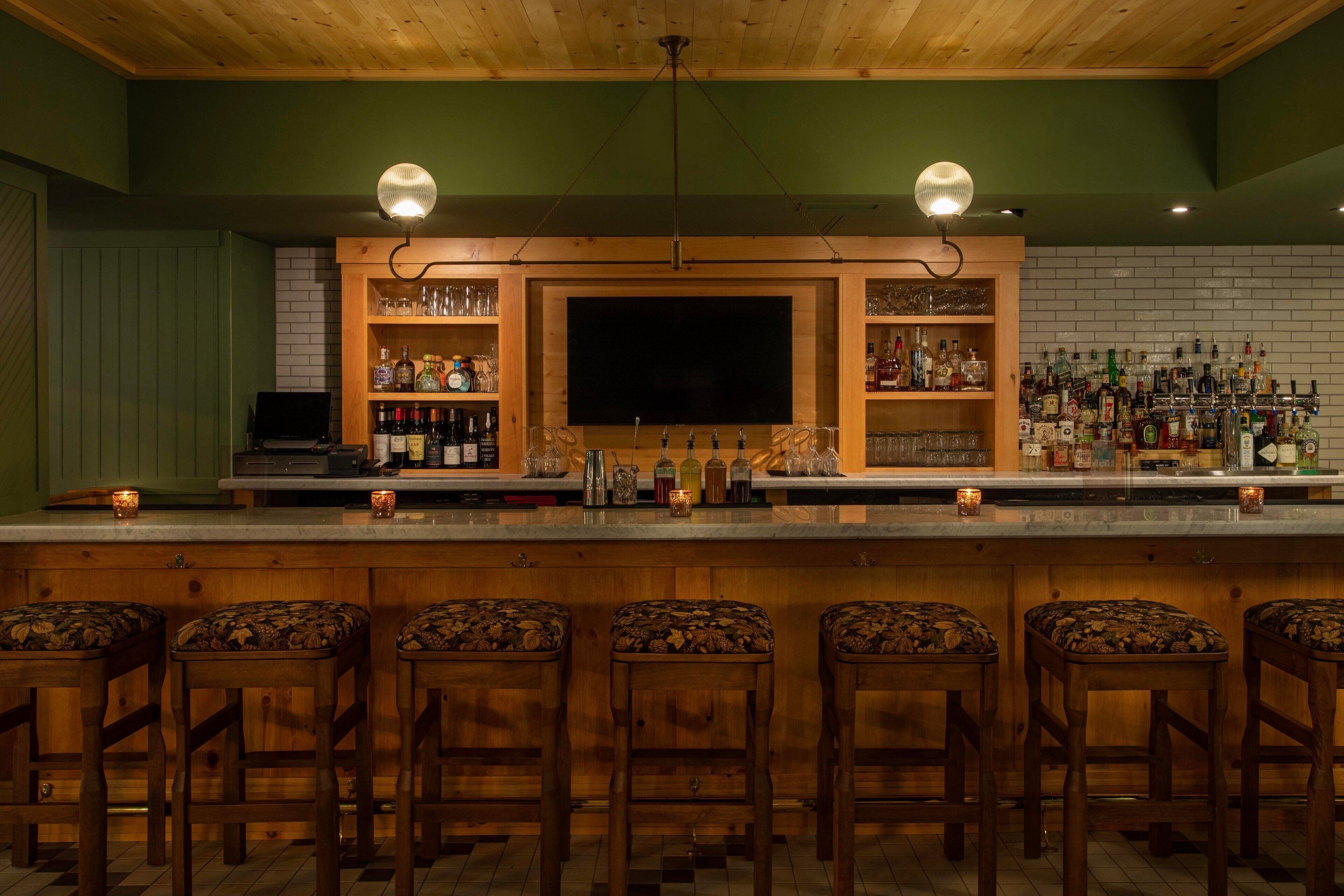 A bar area with wooden stools, a countertop with bottles and glasses, and a television screen.