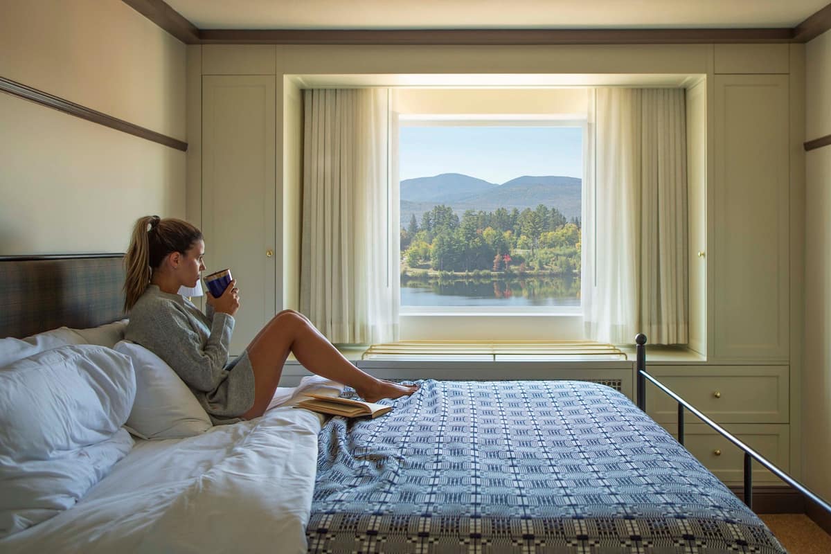 Une chambre avec une grande fenêtre, un lit recouvert d’une courtepointe à motifs, et une vue sur un lac et des montagnes.