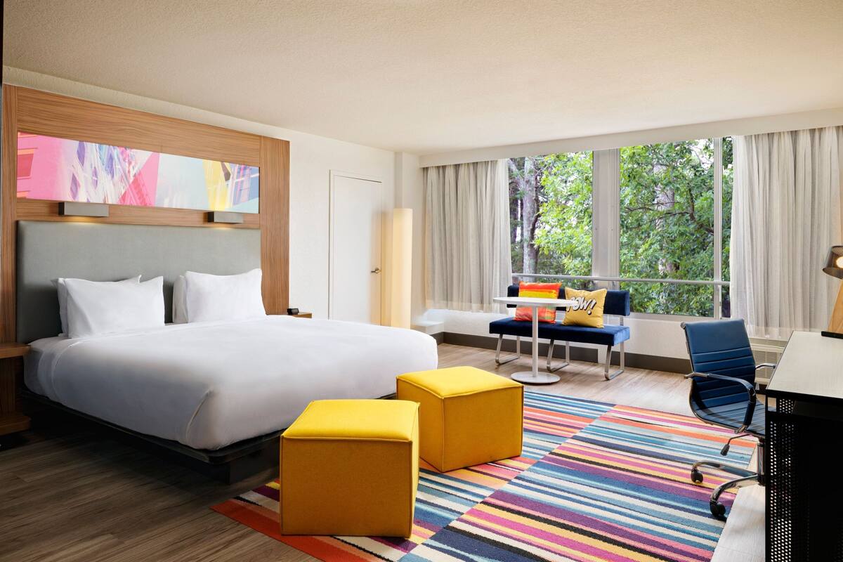 Ein Hotelzimmer mit einem großen Bett, zwei gelben Hocker, einem Schreibtisch und einem Stuhl.