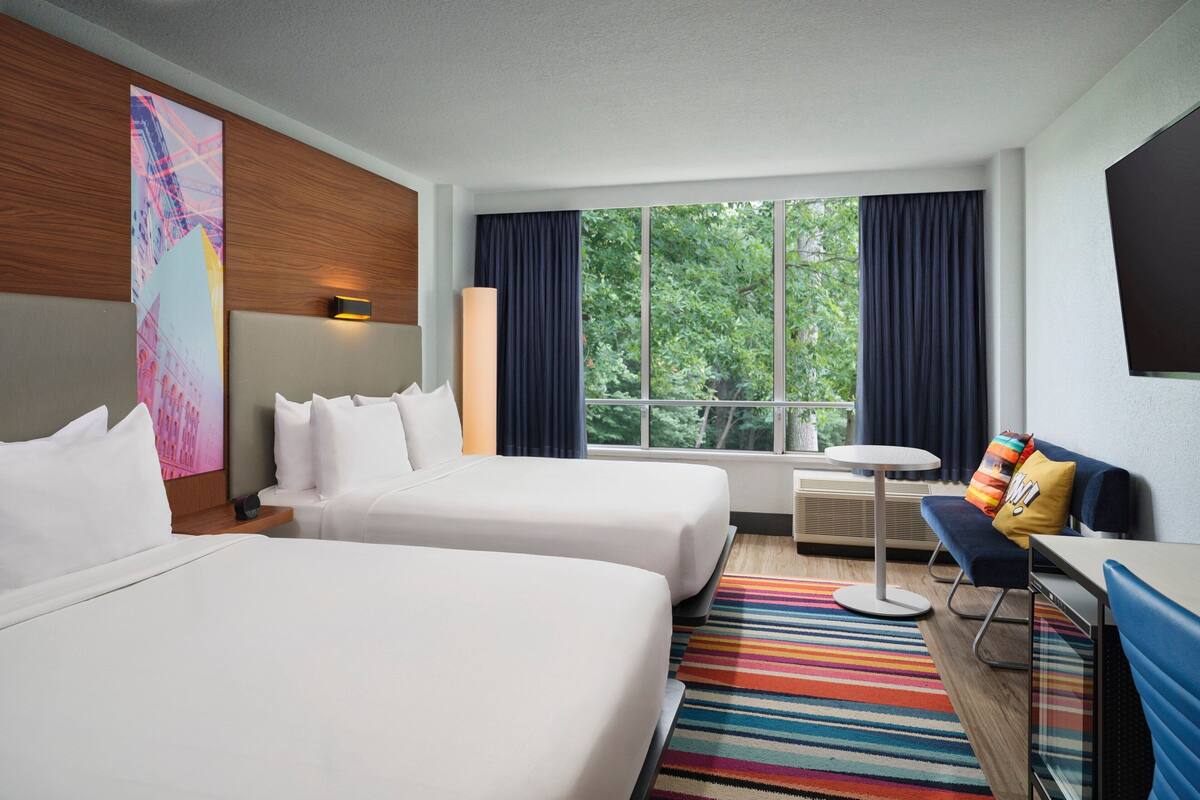 Ein Hotelzimmer mit zwei Betten, einem Schreibtisch, einem Stuhl, einem großen Fenster mit Vorhängen und einem farbenfrohen Streifen Teppich.