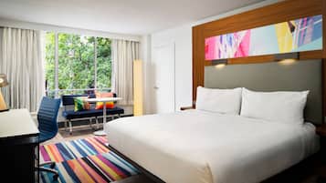 A hotel room with a large bed, a desk with a chair, a colorful striped rug, and a window with curtains.