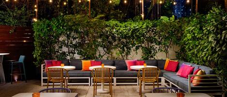 Un área de estar al aire libre con sofá, sillas y mesas, rodeada de vegetación e iluminación de luces colgantes.