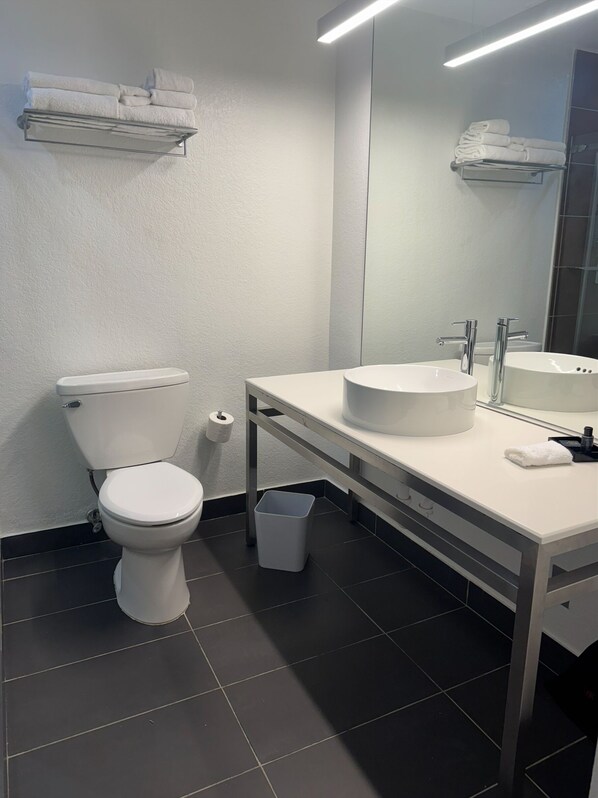 A modern bathroom with a toilet, sink, and mirror. There are towels on a shelf above the toilet and a trash bin next to the sink.