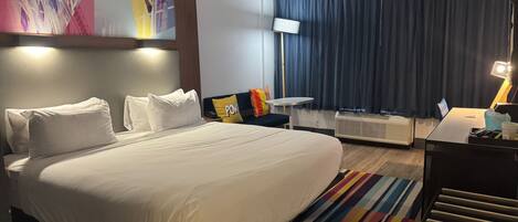 A hotel room with a large bed, a desk, a chair, a small table, a lamp, a heater, and a colorful rug.