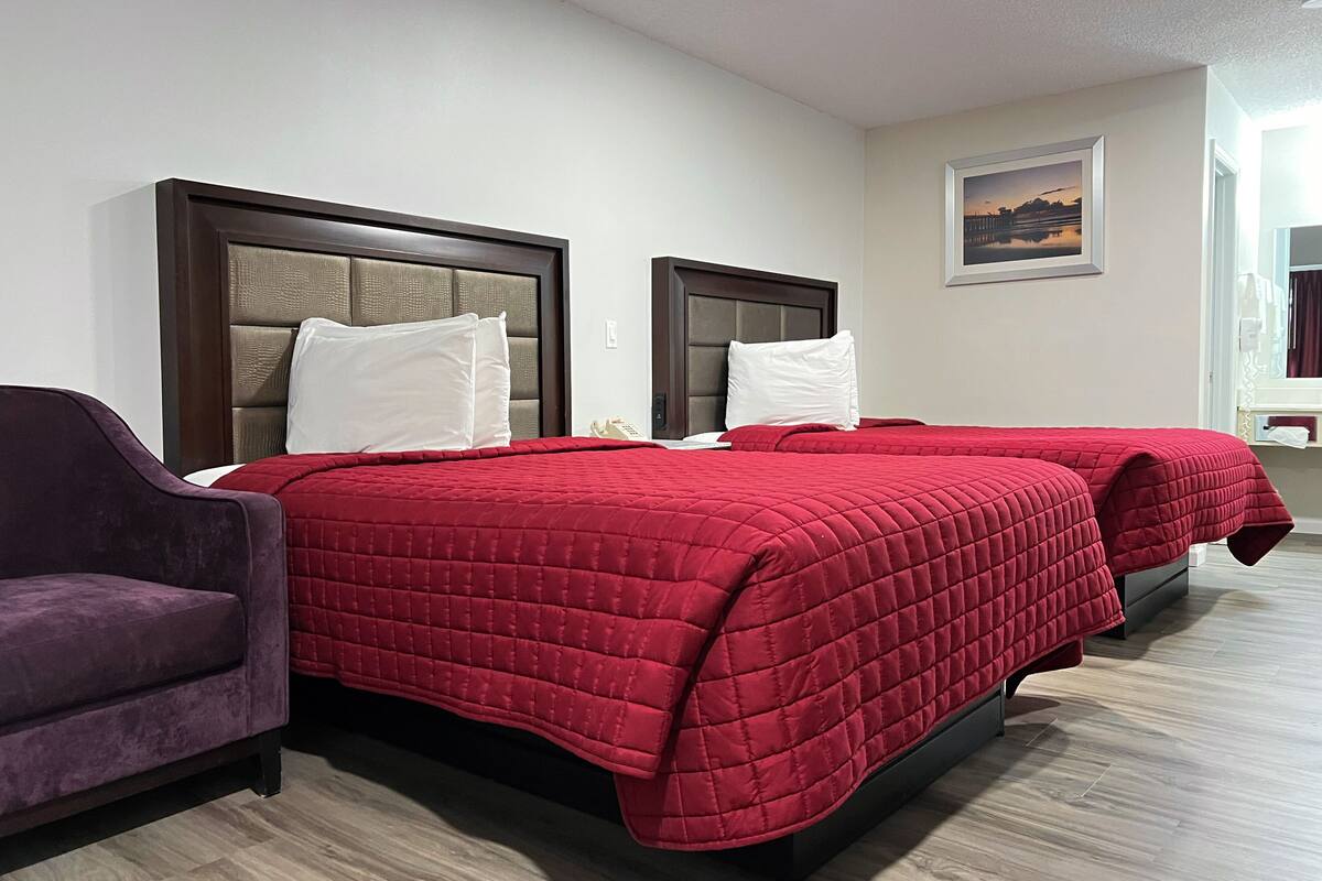 A hotel room with two beds, each with a red quilted bedspread and white pillows. There is a purple armchair to the left of the beds. The room has a wooden floor and a framed picture on the wall.