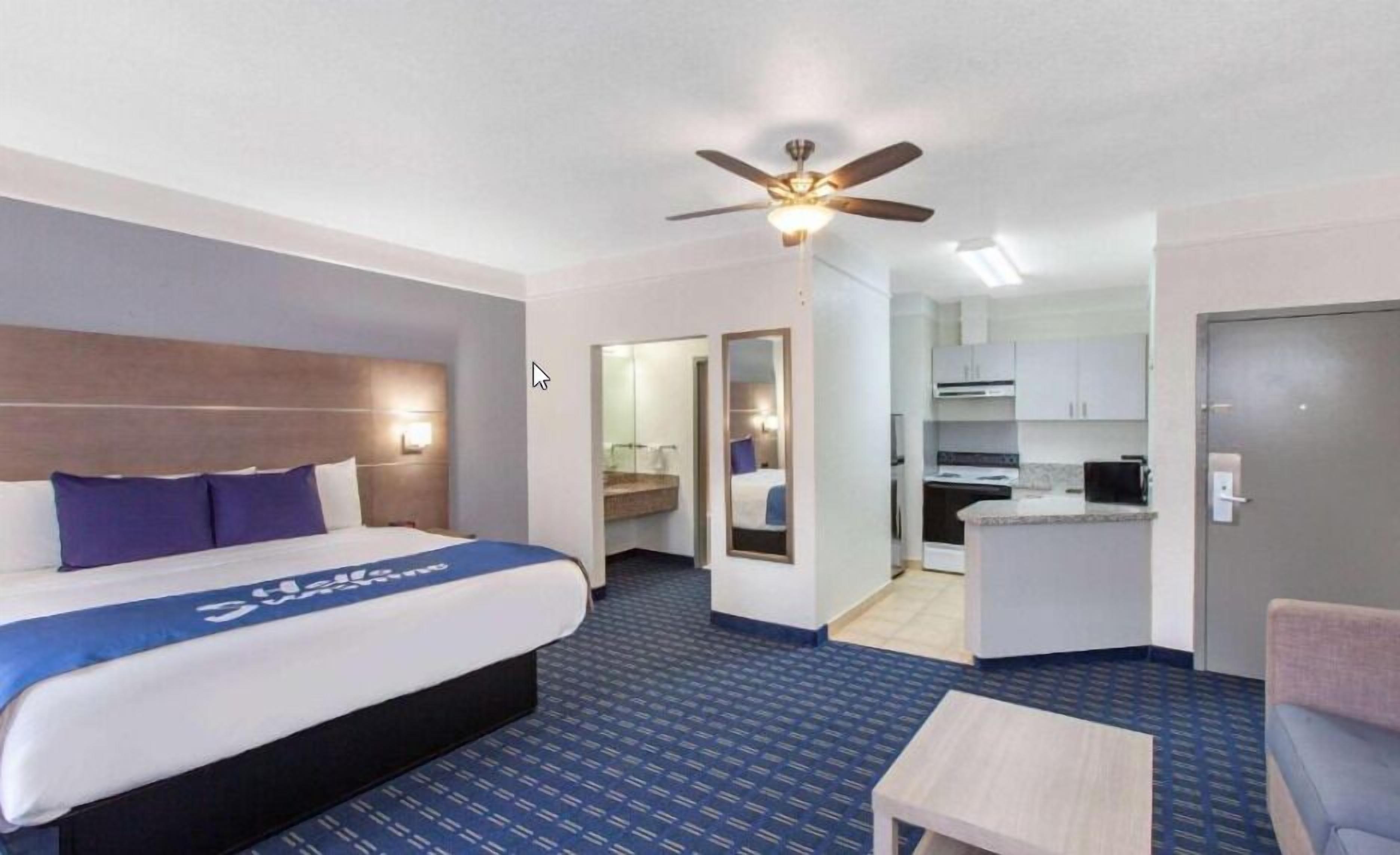 A hotel room with a bed, a desk, a chair, a ceiling fan, and a kitchenette.
