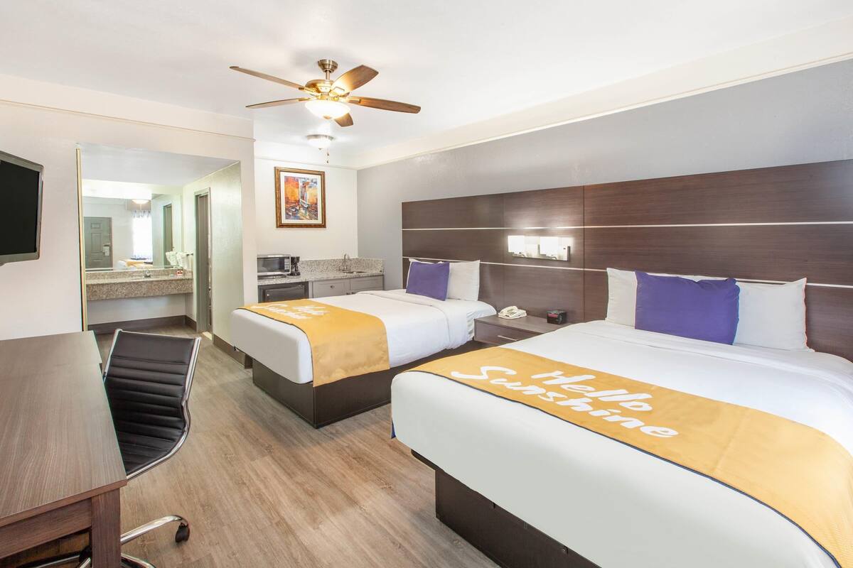 A hotel room with two beds, a desk, a chair, a ceiling fan, a microwave, and a painting on the wall.