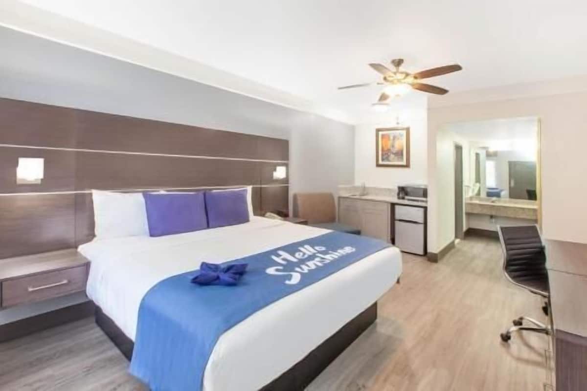 A hotel room with a bed, a desk, a chair, a ceiling fan, and a bathroom mirror.